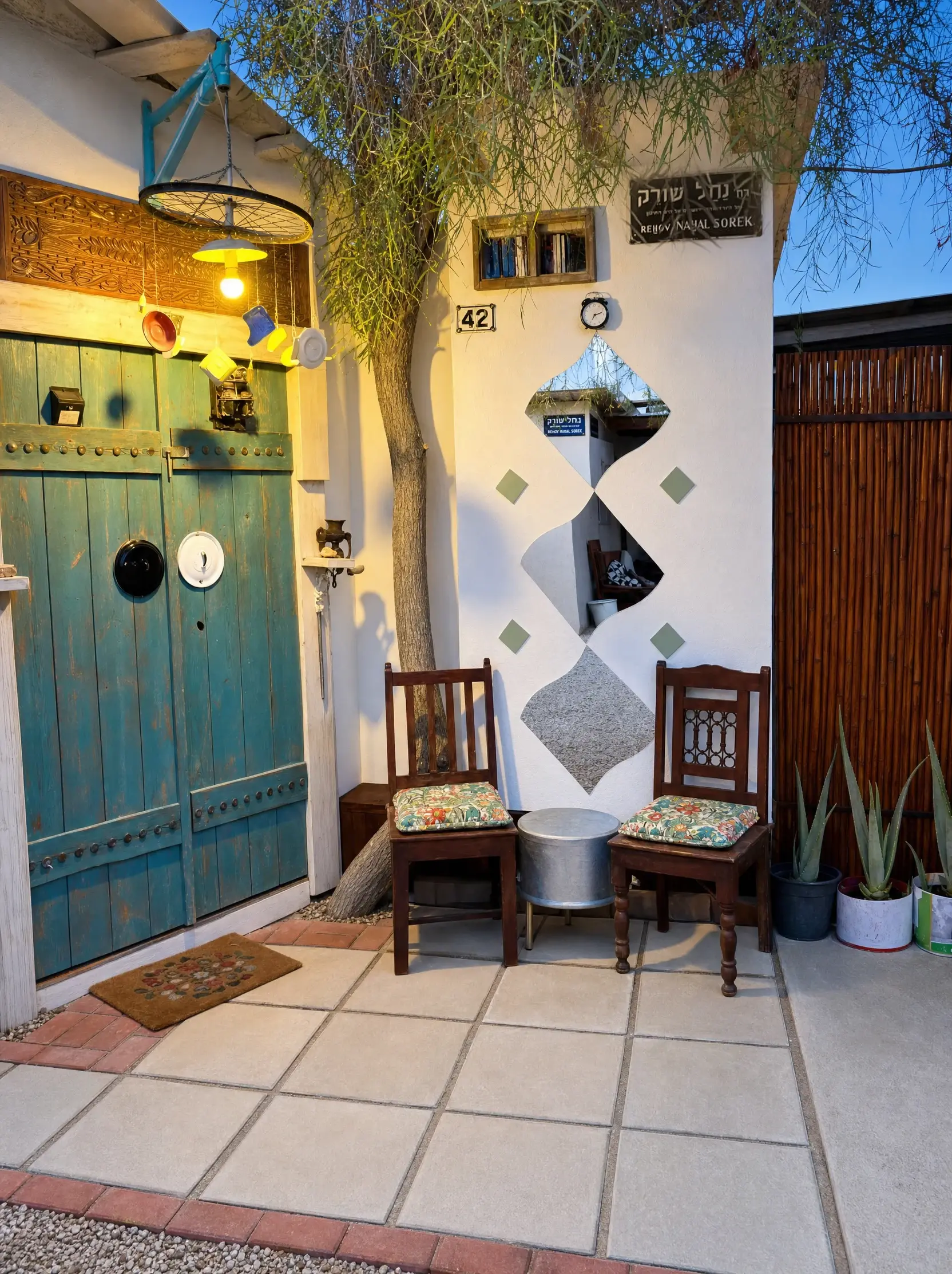 The Eden entrance at dusk: turquoise doors, a bicycle-wheel lamp, a mirror wall and two wooden chairs